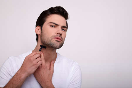Handsome young man isolated. Portrait of a muscular man in a white T-shirt stands on a white background with a black razor in his hand while shaving. The concept of caring for men. Copy spaceの写真素材