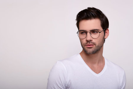 An elegant young white guy with dark hair and a beard in a white T-shirt glasses for vision looks...