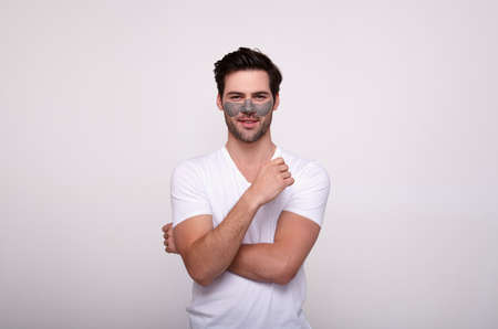 Image of a pensive caucasian male model standing with a clay mask on her face, doing cosmetic procedures at home, isolated over a white wall. Cosmetic advertising. Skin care concept.の写真素材