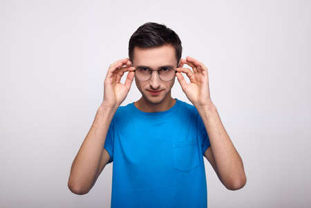 A handsome handsome white man with dark hair and round glasses and a blue T-shirt stares straight, holding on to his glasses. Hypnotic look. A cute young man in a blue T-shirt on a white background.の写真素材