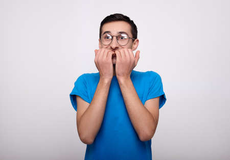 A discouraged young man is very surprised, worried, holds his hands near his face, bites his nails. The young man in a blue T-shirt and glasses experiences strong emotions, looks to the side.の写真素材