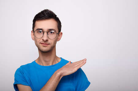 Young stylish, fashionable young guy with trendy haircut in round glasses for vision and a blue T-shirt stands straight, looks into the frame, hand casually swipes dust from the shoulder. Copy space.の写真素材