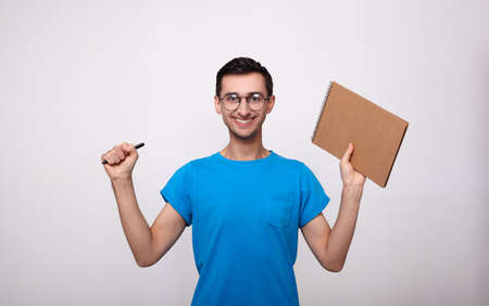 A young cheerful happy guy in a blue T-shirt and round glasses, smiles a broad white smile, holds a notebook and a pen. The atmosphere of joy and victory passed the exam.の写真素材