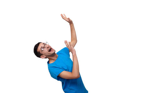 A handsome young man with dark hair with green eyes, in a blue casual T-shirt and round glasses, shouts open his mouth and raised both hands up. Hilarious man is swelled against a white background.の写真素材