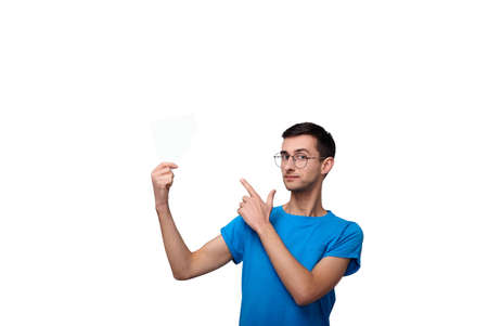 A cute young guy in round glasses and a blue T-shirt in one hand holds an empty white sheet, and the second hand shows at him. Photos of a student on a white background. Copy space.の写真素材