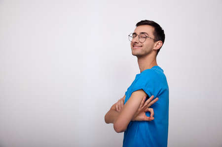 Cute joyful happy young guy with dark hair with glasses and a blue T-shirt stands sideways, folded his hands on his chest and shows the stares the sign Okey, looks at the camera. Copy space.の写真素材