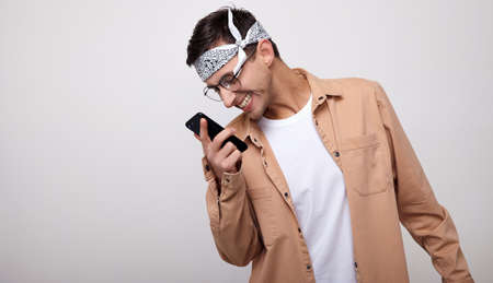 Image of a joyful guy wearing a white t-shirt and bandana, a beige shirt and round glasses for sight, singing and listening to music with a cellphone isolated on a white wall. Copy space.の写真素材