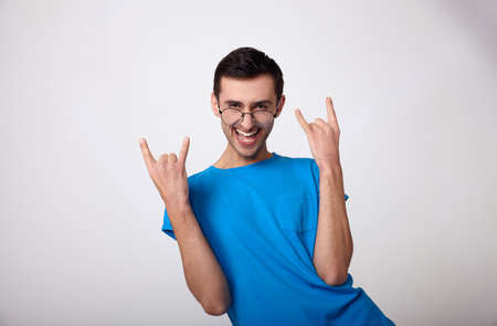 Hilariously happy young man with white skin and dark hair in a blue T-shirt and round glasses shows signs with his hands, opened his mouth and laughs. A man his arms to the side on a white background.の写真素材