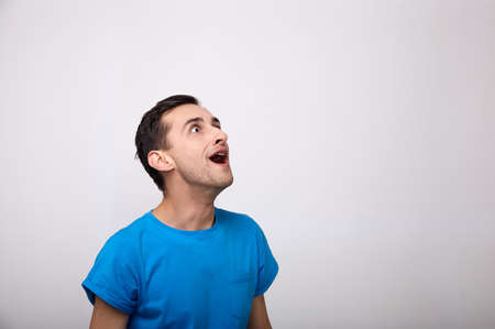 Portrait of a young surprised attractive guy wears a blank blue t-shirt, hears incredible news, stands on a white background with copy space, his mouth wide open in shocked expression. Copy space.の写真素材