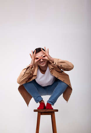 A young white man in stylish fashionable clothes, red socks, squatting on a high wooden chair and makes glasses with his hands. A cheerful student has fun.の写真素材