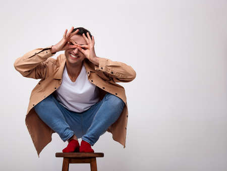Joyful positive young man is sitting on a chair and smiling while in a good mood. A young white man in stylish fashionable clothes, red socks, squatting on a high wooden chair. Copy space.の写真素材
