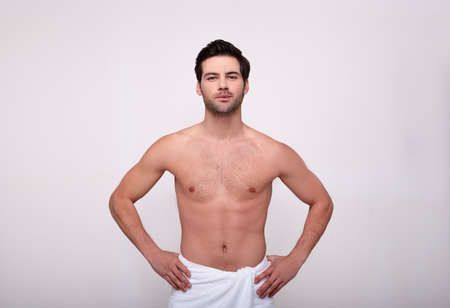 The charismatic brunette with brown eyes, clean skin, stands half-naked in a white towel on his hips, his hands rested in the sides, looks straight.の写真素材