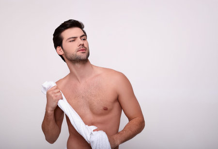 Adorable nice confident athletic white man with dark eyes after shower, shirtless, wipes body with a white towel, looks to the side. He is after a morning shower, isolated on a white background.の写真素材