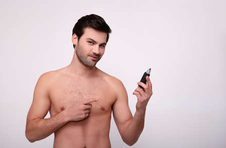 The cute attractive smiling guy looks at the camera, points his finger at the trimmer to remove the hair in his hand. The concept of male hygiene, body care, hair removal. Copy space.の写真素材
