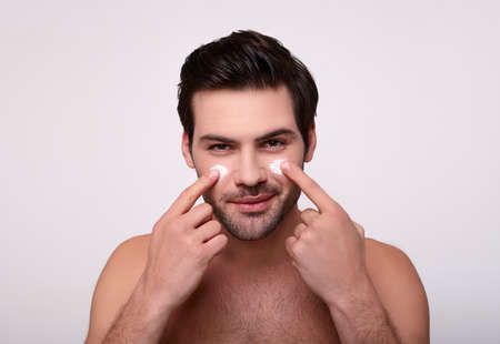 Skin care. Lovely caucasian shirtless man applying cream on his face and looking at himself with a smile while standing over gray background and looking up. Close-up Space for text.の写真素材