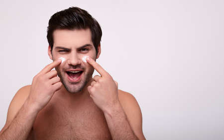 Skin care. Handsome young shirtless man applying cream at his face and looking at himself with smile while standing over gray background and looking up. Close-Up. Space for text. Copy space.の写真素材