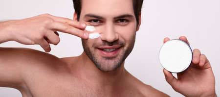 Close-up portrait of smiling shirtless young man applying facial cream, isolated on gray background. Skin care concept.の写真素材