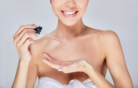The clipped frame, an image of a young girl with a white smile, stands in a white towel. holds the oil for massage. Skin hydration, body care, healthy skin, spa treatments. Photo on a gray background.の写真素材