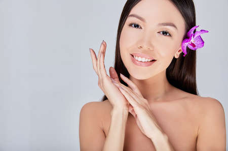 A young cute girl Asian with clean healthy skin, bare shoulders, with a purple flower orchid in her hair, folded her hands near her face and smiles gently, looks straight. Copy space.の写真素材