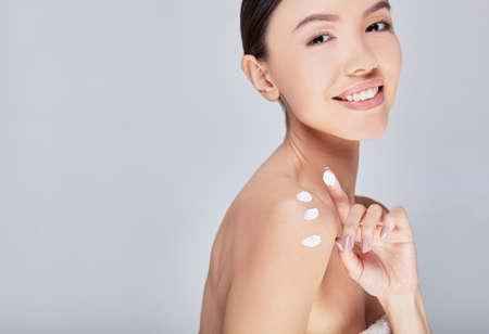 Image of a young gentle Asian woman with perfect healthy skin, the lady came out of the shower and applied a finger moisturizing cream on her shoulder, posing against a gray background. Copy space.の写真素材