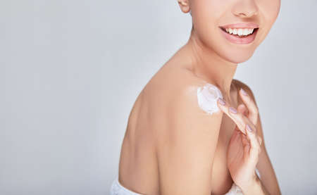 Close-up, a young Asian woman with beautiful healthy skin is sitting on a fucking background, applying moisturizer to her shoulder. The concept of self-care, facial hydration. Copy space.の写真素材