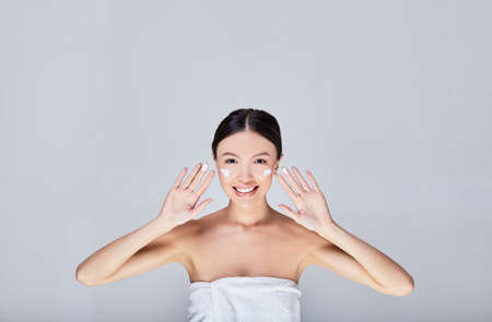 Beautiful young woman applying anti-wrinkle cosmetic cream to moisturize. Cute Asian woman with cream on her face, treatment on her face. Photo isolated on a light gray background.の写真素材
