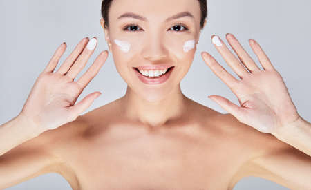 Close-up, a young beautiful happy Asian woman with dark hair and brown eyes, with a white smile, puts cream on her face with her fingers. hoto isolated on a light gray background.の写真素材