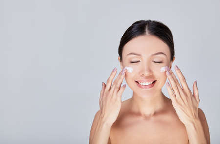 Beautiful young woman applying cosmetic cream. Cute Asian woman with cream on her face, treatment on her face. Photo isolated on a light gray background. Copy space.の写真素材