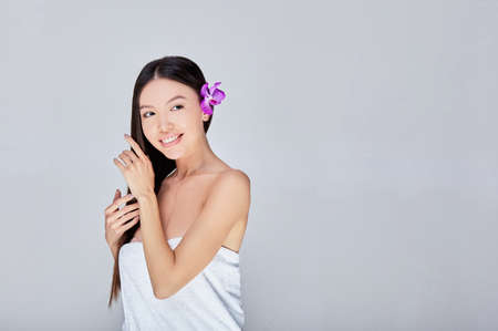 A pretty romantic young Azitian lady with long dark thick hair and a flower behind her eye, stands in a white half-shade and touches her hair with her hands, smiles, looks to the side. Copy space.の写真素材