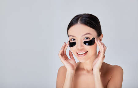 Beautiful young mixed race woman caring for skin, smiling, applying hydrogel patches for restoration under her eyes, looking in the mirror, collagen pads for a beautiful concept of facial skin care.の写真素材
