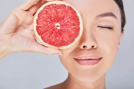 Close-up portrait of a young beautiful Asian woman with perfect skin, dreamily closed her eyes, holds a grapefruit near her face. Photo on a gray background. The concept of cosmetics with vmitamine C.の写真素材