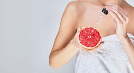 The cropped frame, a young white girl with beautiful healthy skin, applies pipette oil on a cut grapefruit. Vitamin C Cosmetics Concept. Copy space.の写真素材