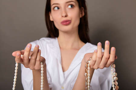 A young beautiful white lady with long dark hair, in a white shirt, holding pearls in her hand, thinking about something, making a choice. The concept of natural cosmetics, pearl extract.の写真素材