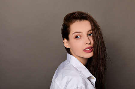 Cute young Caucasian girl with blue eyes, with long dark wet loose hair in a white shirt posing standing on a dark background, looking to the side. Copy space.の写真素材