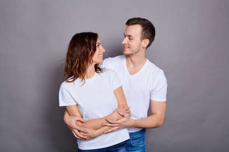 Portrait of two young, handsome, attractive, cheerful, positive couple, people hugging laughing, wearing white basic t-shirts, looking at each other, isolated over gray background. Copy space.の写真素材