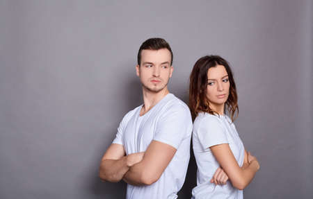 A young couple in white base T-shirts, standing with their backs to each other, hands folded on their chests, looking in, the couple depressed by quarrel, posing against a gray background. Copy space.の写真素材