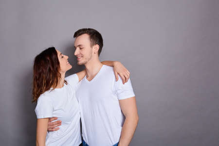 Portrait of a Caucasian lovely couple - a man in a white T-shirt hugs his charming, cute woman in a white T-shirt and they both look at each other, standing on a gray background. Copy space.の写真素材