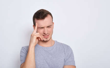 Close-up of a young man isolated on a white background in a gray casual t-shirt touching his temples with his fingers, as if suffering from a severe headache. Headache concept. Copy space.の写真素材