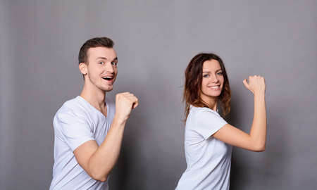 Young cute cheerful couple, in white basic t-shirts, smiling, posing together at the camera while working or jumping, running, standing on gray background.の写真素材