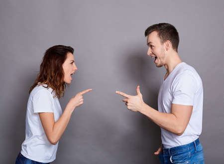 Photo of an annoyed aggressive Caucasian couple in basic white T-shirts screaming at each other, standing face to face, isolated on a gray background. Young lovers quarrel, elevate relationships.の写真素材