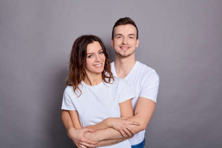 Portrait of two young, handsome, attractive, cheerful, positive couple, people cuddling laughing, wearing white basic t-shirts, isolated over gray background. Copy space.の写真素材