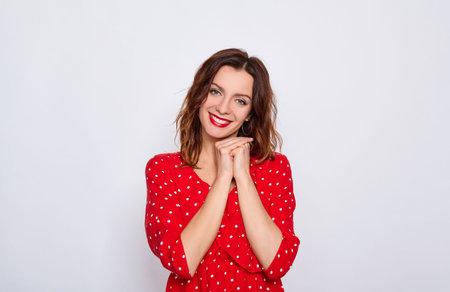 Charming cute young brown-haired woman with blue eyes and bright makeup, in a red dress in a white dot, folded her hands under the chin, smiles and looks straight posing on a white background.の写真素材