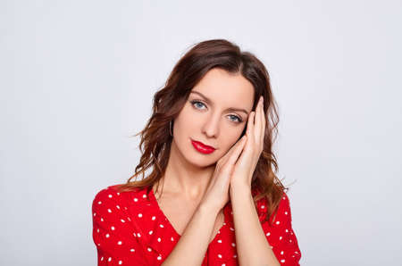 Image of a pretty young woman posing with blue eyes and red hair, in a red dress, isolated over a white wall background with bright red lipstick, folded her hands under her cheek and look straight.の写真素材