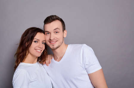 Portrait of a Caucasian lovely couple - a man with blue eyes hugs his charming, sweet woman, and they both look at the camera, standing against a gray background. Copy space.の写真素材