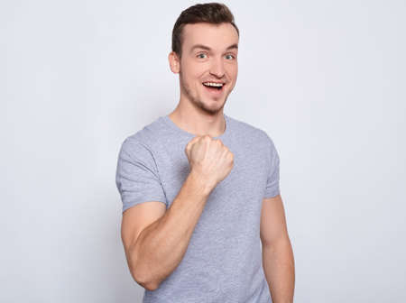 A handsome handsome young Caucasian attractive man with a wide snow-white smile, in a gray basic T-shirt raised one fist in a victory gesture, rejoices in victory, good news, good luck.の写真素材