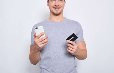 Cropped frame, a young handsome man in a gray T-shirt is thinking about online shopping through the Internet, holding a credit card and a smartphone on a white background.の写真素材