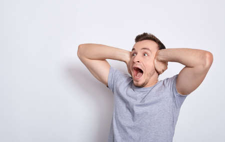 A young screaming angry man in a basic gray T-shirt, screaming noisily at the noisy neighbors from above, covering his ears with both hands, does not want to hear bad news. Copy space.の写真素材