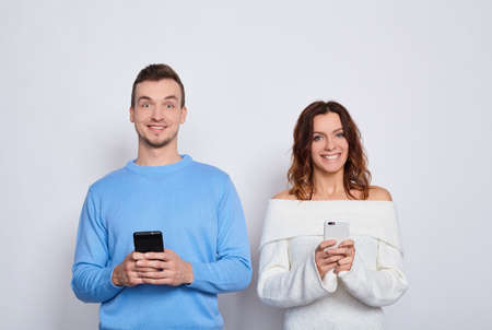Photo of expressive funny cheerful people man and woman in casual light clothes smiling, looking at each other, while both using mobile phones isolated on a white background.の写真素材