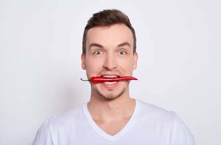 Close-up, surprised pretty young charming pretty white guy with green eyes in a white T-shirt, holds a red chili pepper in his teeth and playfully bites it, isolated on a white background.の写真素材
