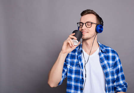 A young happy cheerful joyful man in glasses, in a white T-shirt and blue shirt, holds a black coffee cup, listens to his favorite music with headphones, closed his eyes and smiles with pleasure.の写真素材